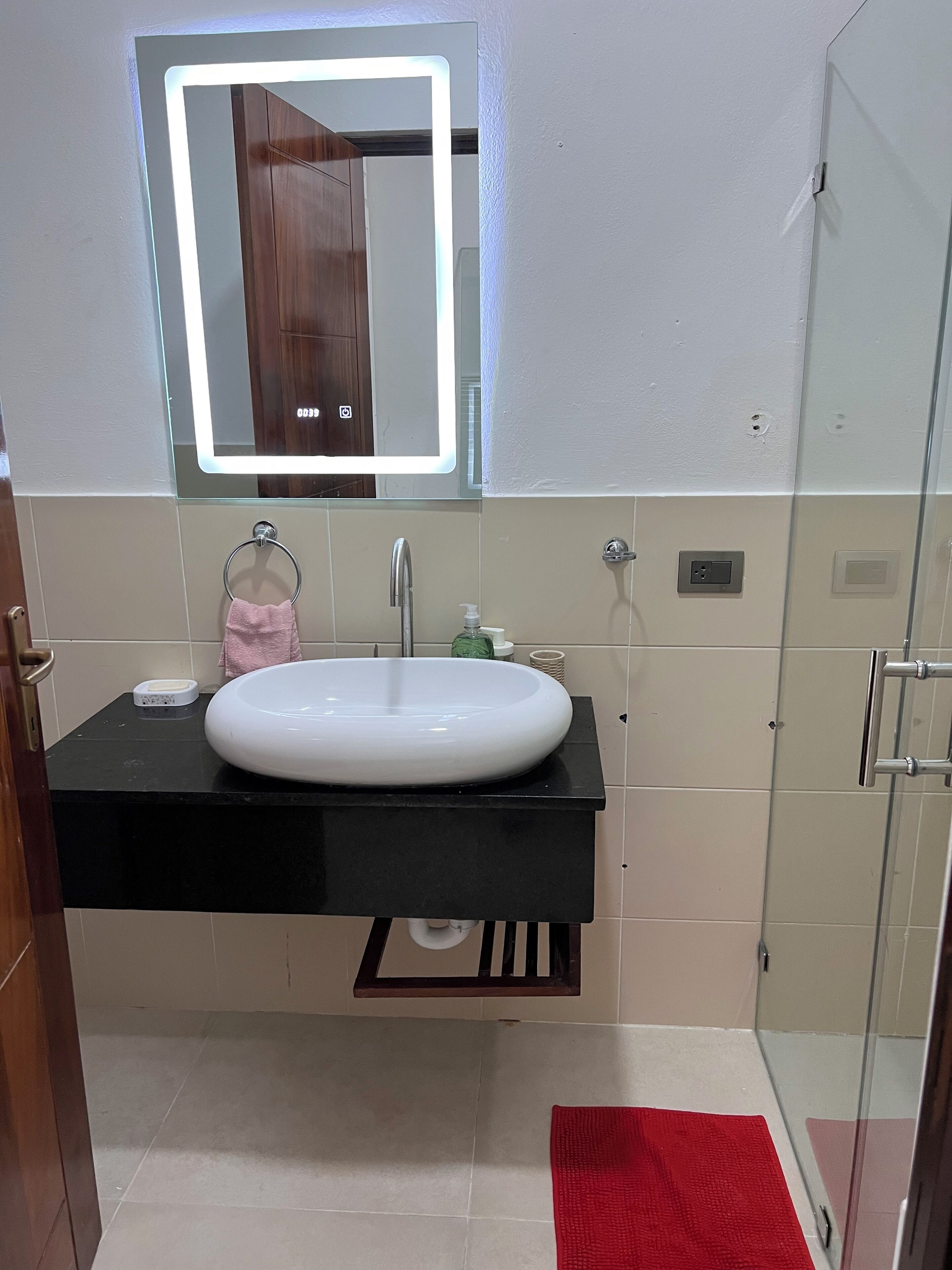 A bathroom with a white sink, a mirror with a light, a towel rack, and a red rug.