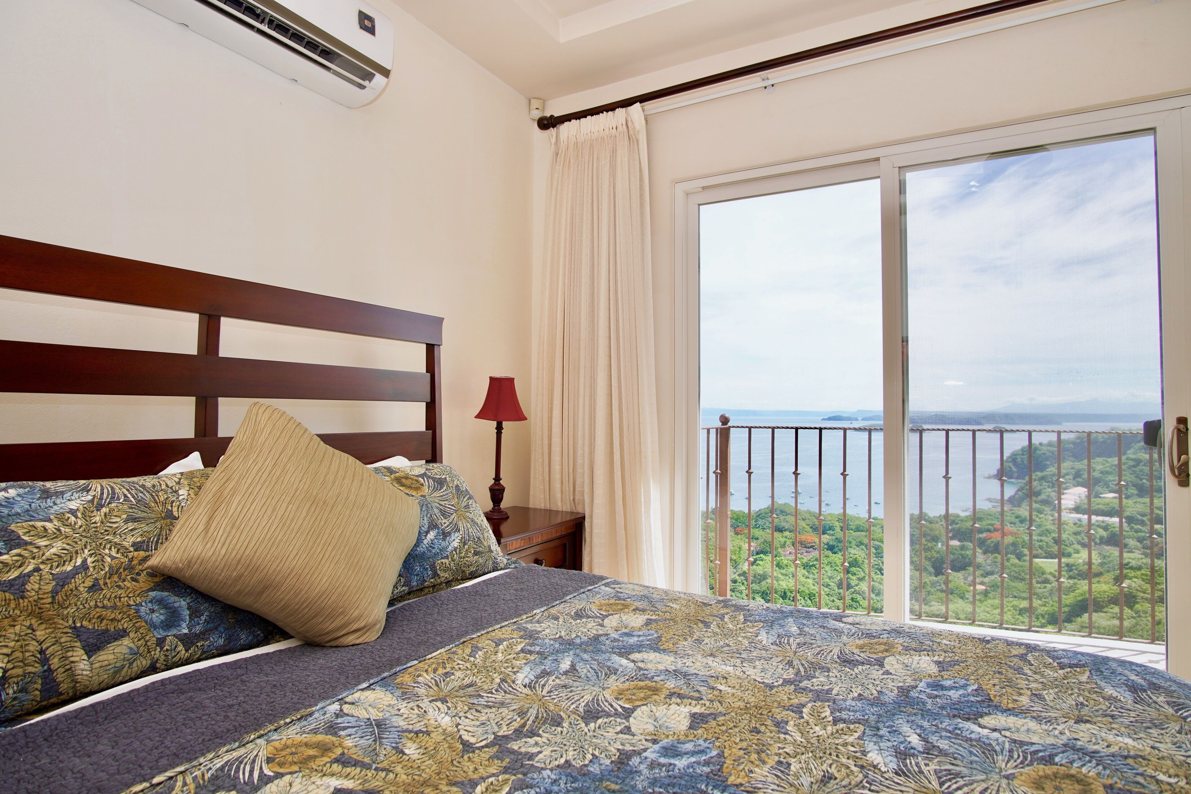 A bedroom with a large bed, a nightstand, a red lamp, and a view of a scenic landscape through a sliding glass door.