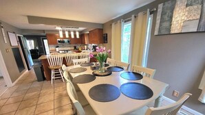 Un comedor con una mesa y sillas blancas, un florero con flores y una cocina al fondo.