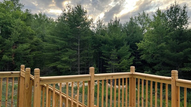 A wooden deck with a railing, surrounded by dense green trees. - Peaceful newly built home between Fish Creek and Ephraim, WI. 3 bed/ 2 bath. (Fish Creek)