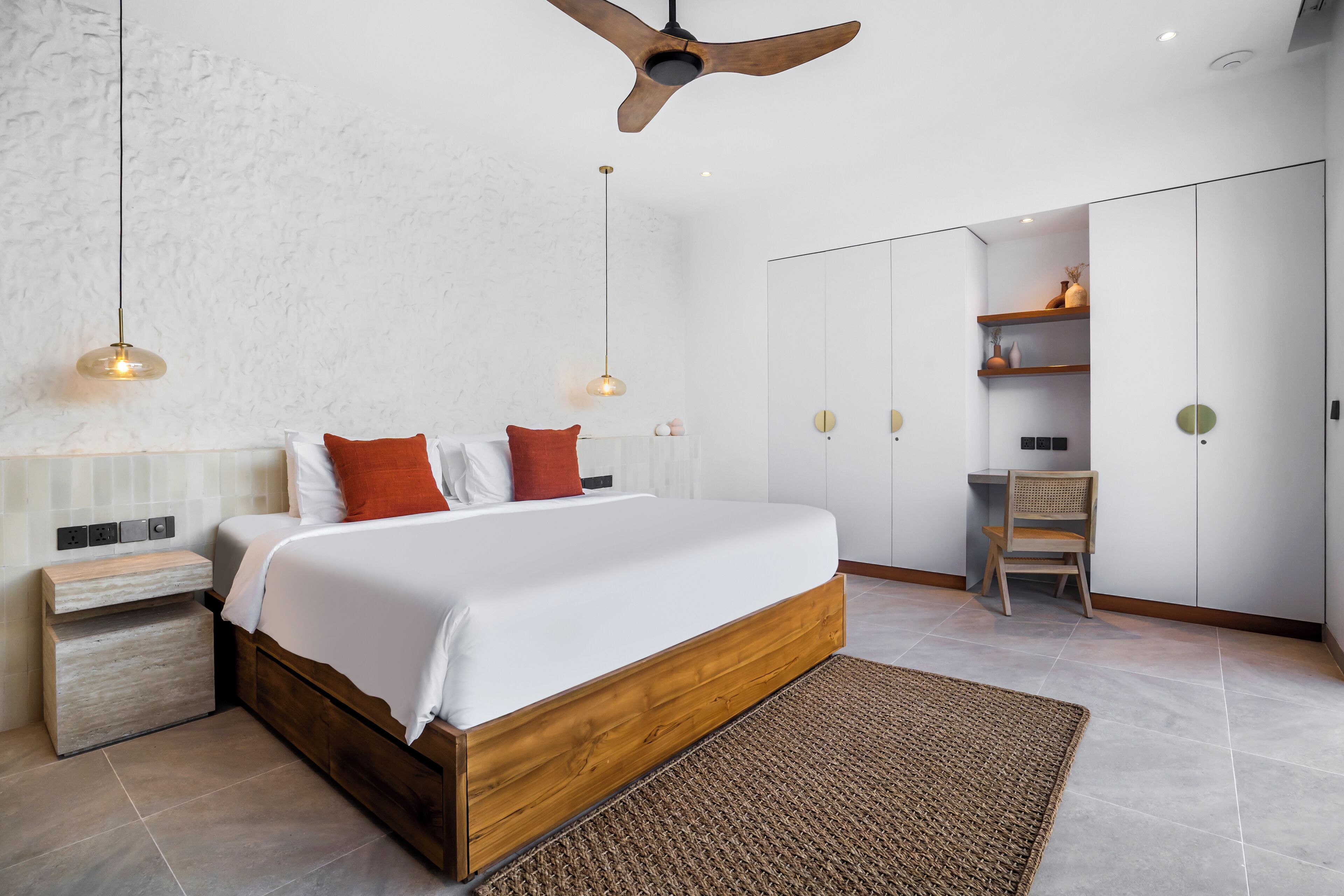 Une chambre avec un lit en bois, du linge de lit blanc, un tapis tissé, un bureau avec une chaise et un ventilateur de plafond.