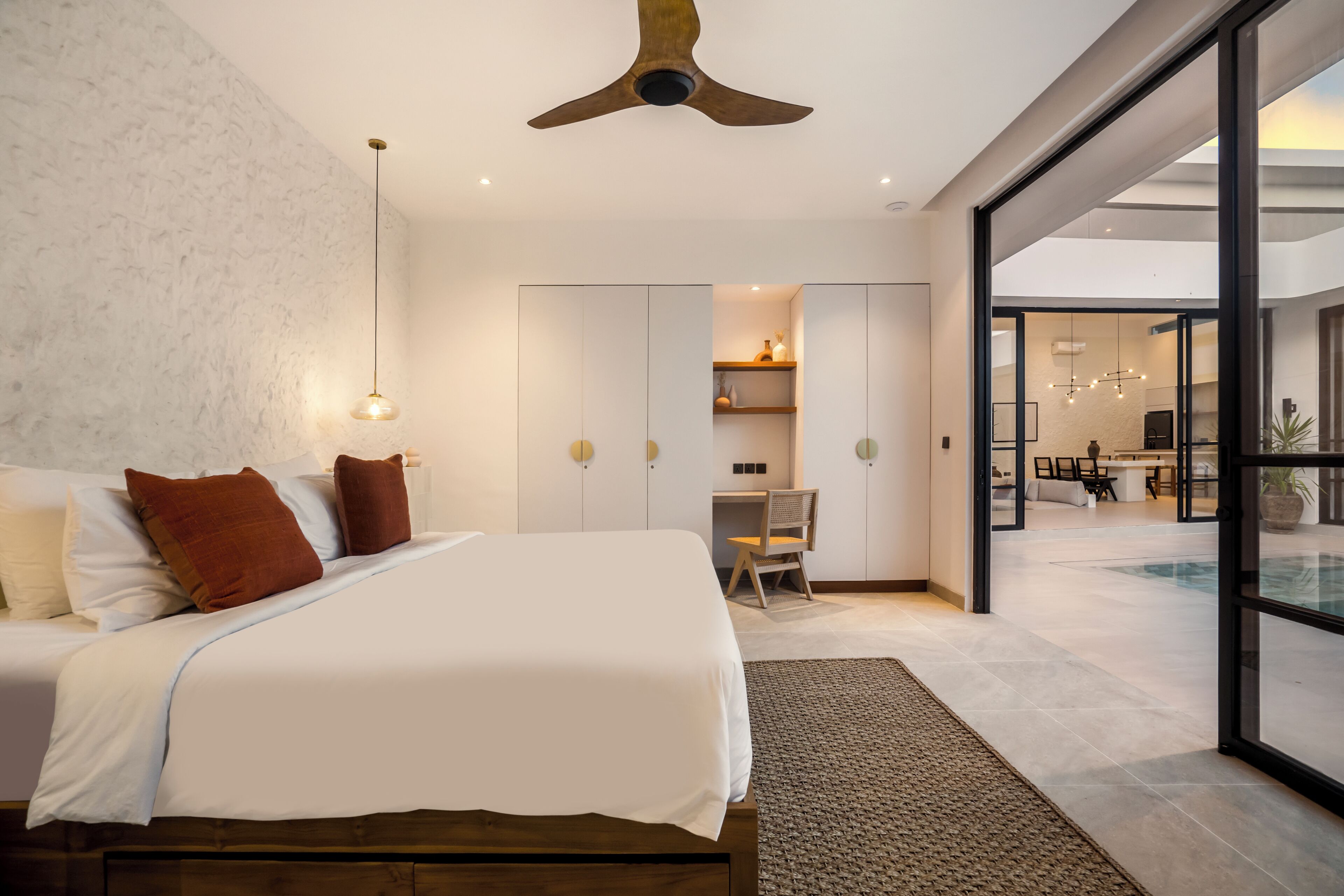 A modern bedroom with a large bed, a ceiling fan, and a view of the living area through a glass door.