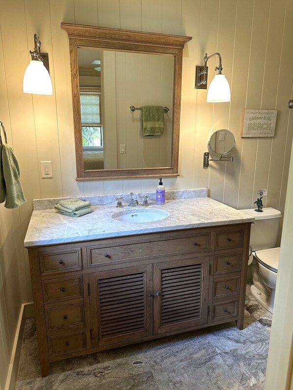 A bathroom with a wooden vanity, a sink, and a large mirror. There are two wall-mounted lights, a towel rack, and a sign that says "Welcome to the Beach." - Admired Poipu beach house in a perfect location, now a vacation home! (Koloa)