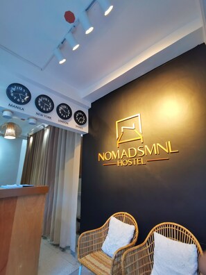 A reception area with a black wall featuring a gold logo for NomadsMNL Hostel. There are clocks displaying times for Manila, New York, London, and Sydney. Two wicker chairs with white cushions are placed in front of the wall. - NomadsMNL Hostel (Parañaque)