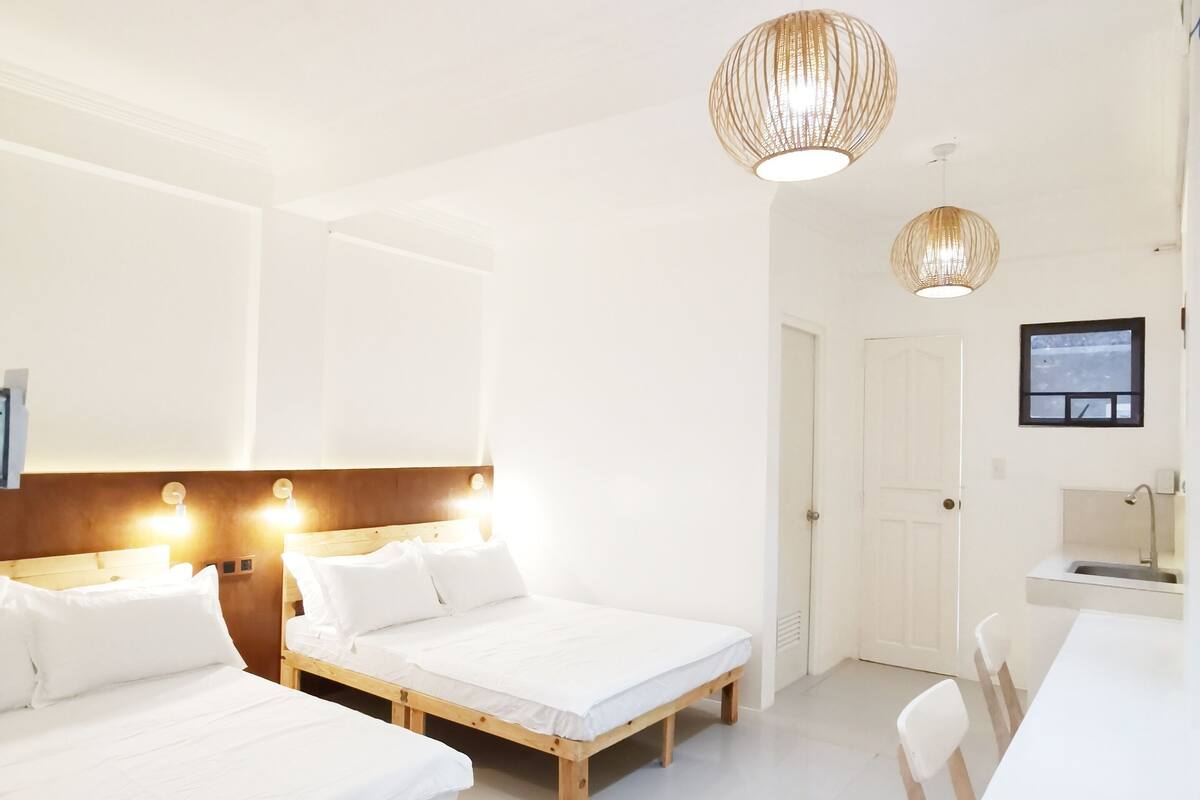 Una habitación de hotel moderna con dos camas individuales, un cabecero de madera, una mesa de comedor y una zona de cocina.