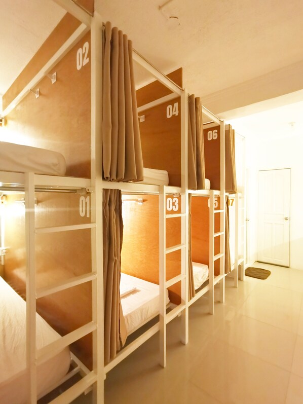 A dormitory-style room with bunk beds numbered 01 to 06, white walls, and a door in the background. - NomadsMNL Hostel (Parañaque)
