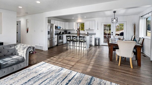 A spacious living area with a grey sofa, a dining table with chairs, a kitchen with white cabinets, and a large window. - Large Home in Quiet Neighborhood (Taylorsville)