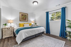 Un dormitorio con cama, mesitas de noche, una ventana con cortinas, una planta y un cuadro en la pared.