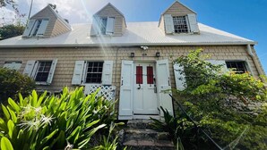 A two-story house with a gabled roof, a central door with red shutters, and a small porch area. - Crucian Fusion: Historic charm meets lush beauty in a rare garden oasis in downtown Christiansted! (Christiansted)