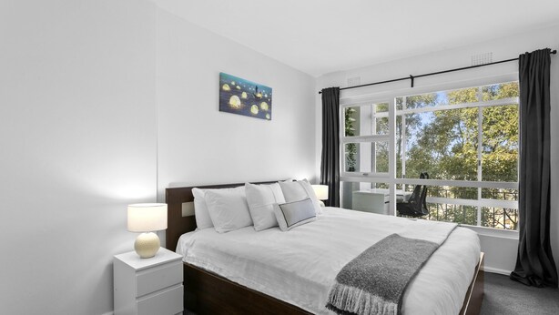 Un dormitorio con una cama grande, una mesita de noche con lámpara, una ventana con cortinas y un cuadro en la pared.