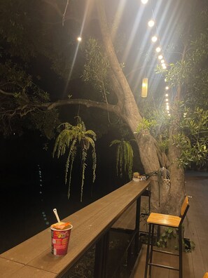 A wooden bench with a can of food and a straw, a hanging light fixture, and a tree with hanging ferns. - Banmai87 Guest House (Ayutthaya)