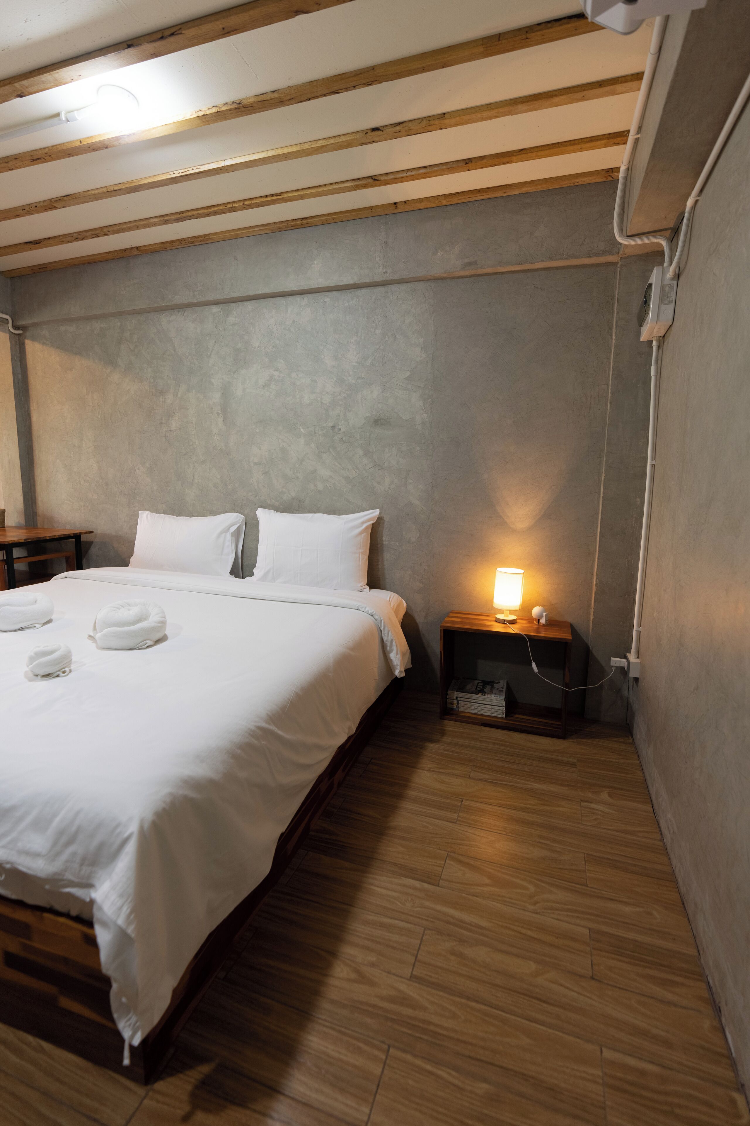 Een keurig opgemaakt bed met witte lakens en kussens, een nachtkastje met een lamp, en een houten vloer.