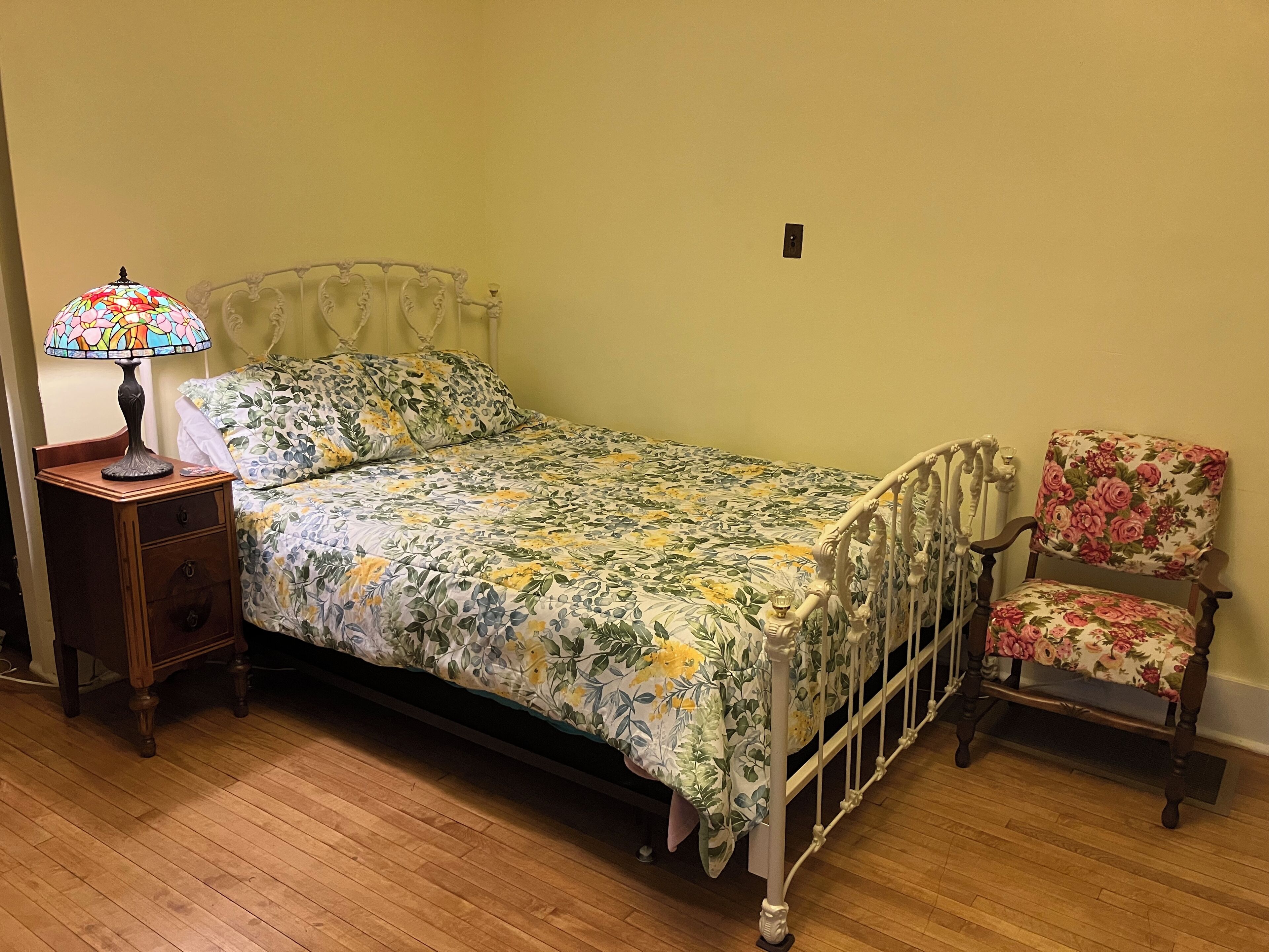 A single bed with a floral bedspread, a nightstand with a lamp, a chair with a floral cushion, and a wooden floor.