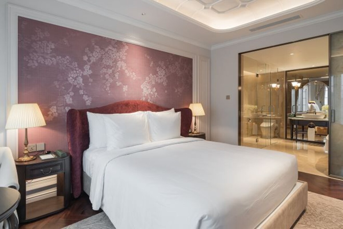 A hotel room with a large bed, two bedside tables with lamps, a red headboard, and a glass-enclosed bathroom.