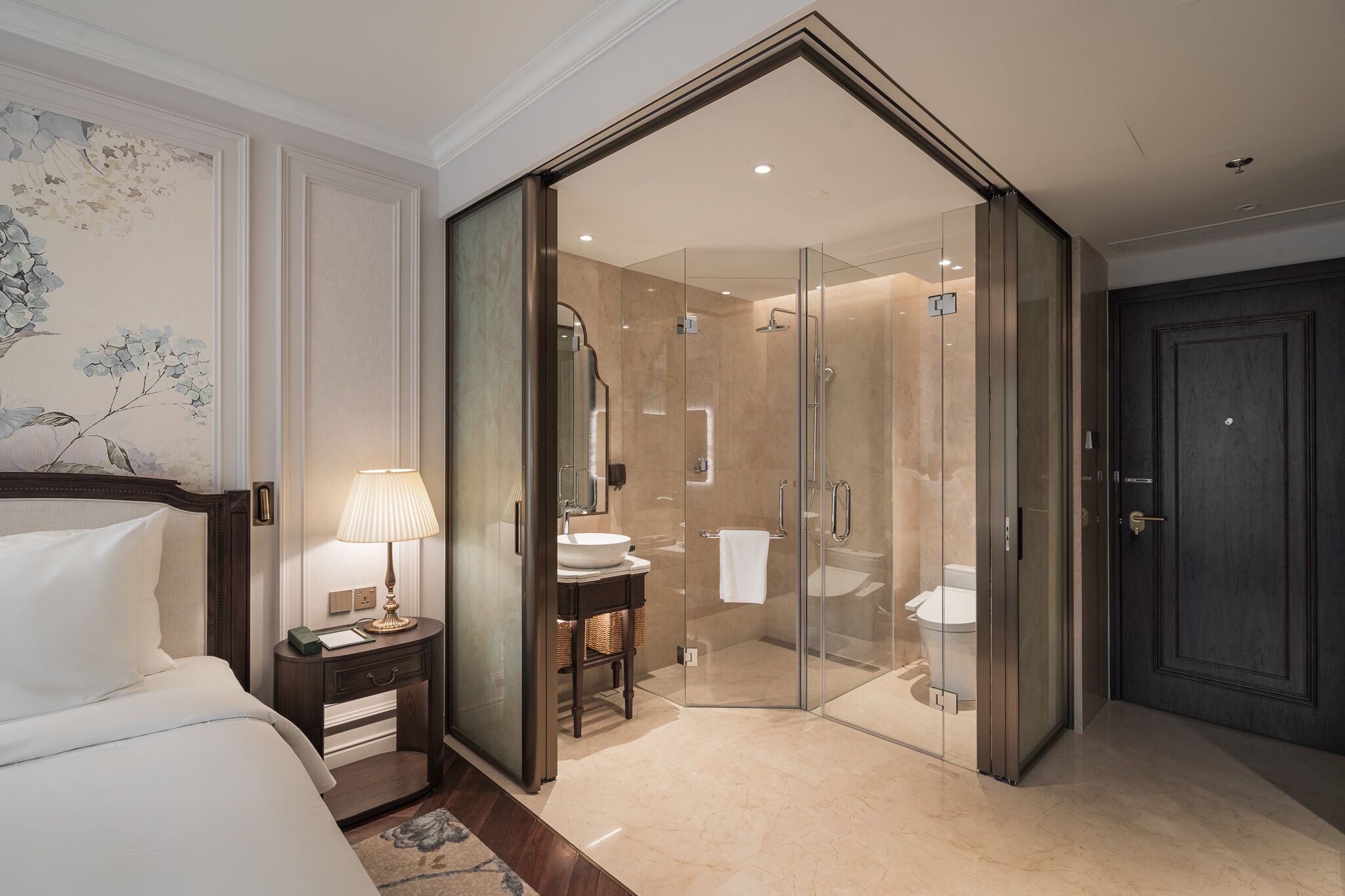 Una habitación de hotel moderna con cama, mesita de noche, lámpara y zona de ducha con mampara de vidrio.
