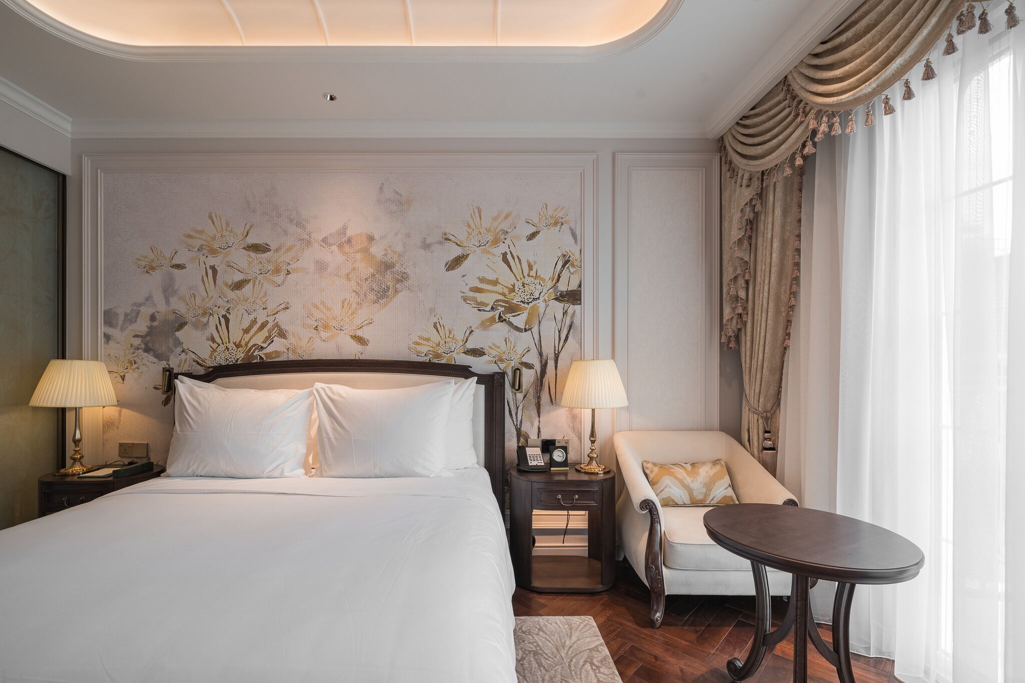 Quarto de hotel com uma cama grande, duas mesas de cabeceira com luminárias, uma mesinha redonda, um sofá e um mural floral decorativo na parede.