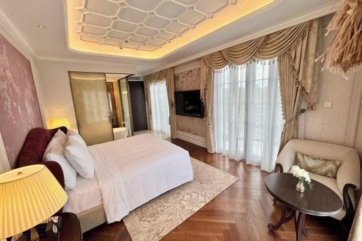 A hotel room with a large bed, a sitting area with a table, a TV mounted on the wall, and a decorative ceiling.
