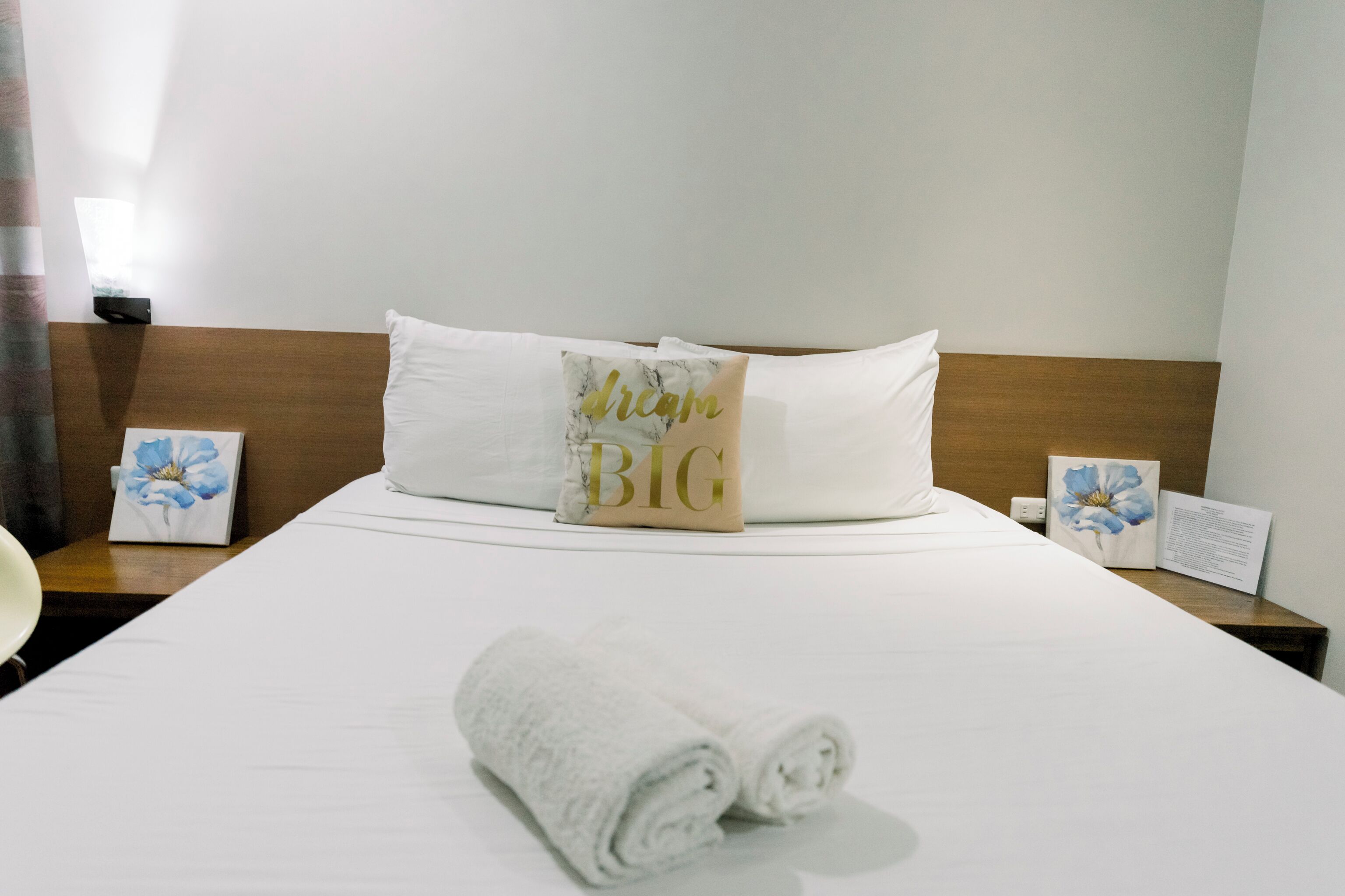 A neatly made bed with white linens, a rolled towel, and a pillow with 'dream BIG' text.