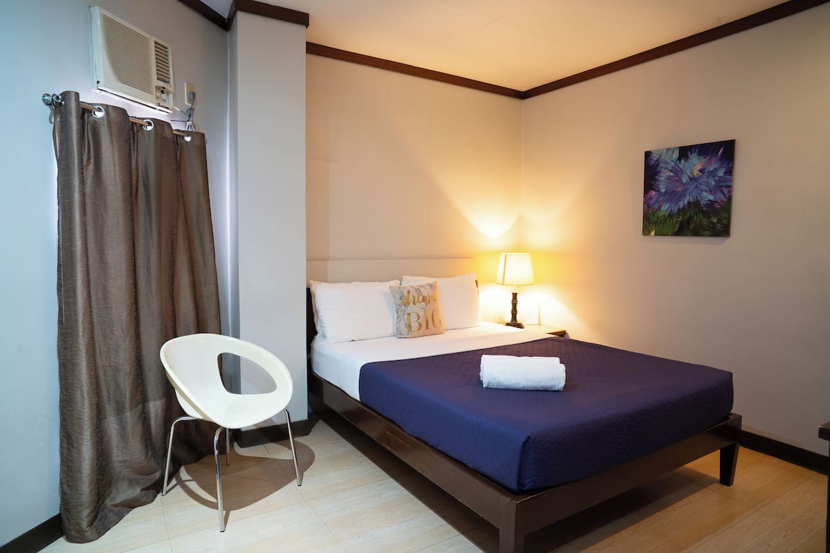 A hotel room with a bed, a chair, a lamp, a painting, and an air conditioning unit.