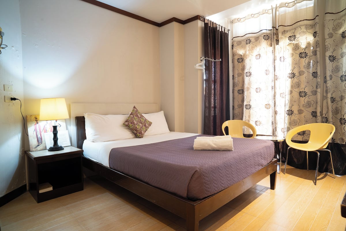 A bedroom with a bed, a nightstand with a lamp, a chair, and a window with curtains.