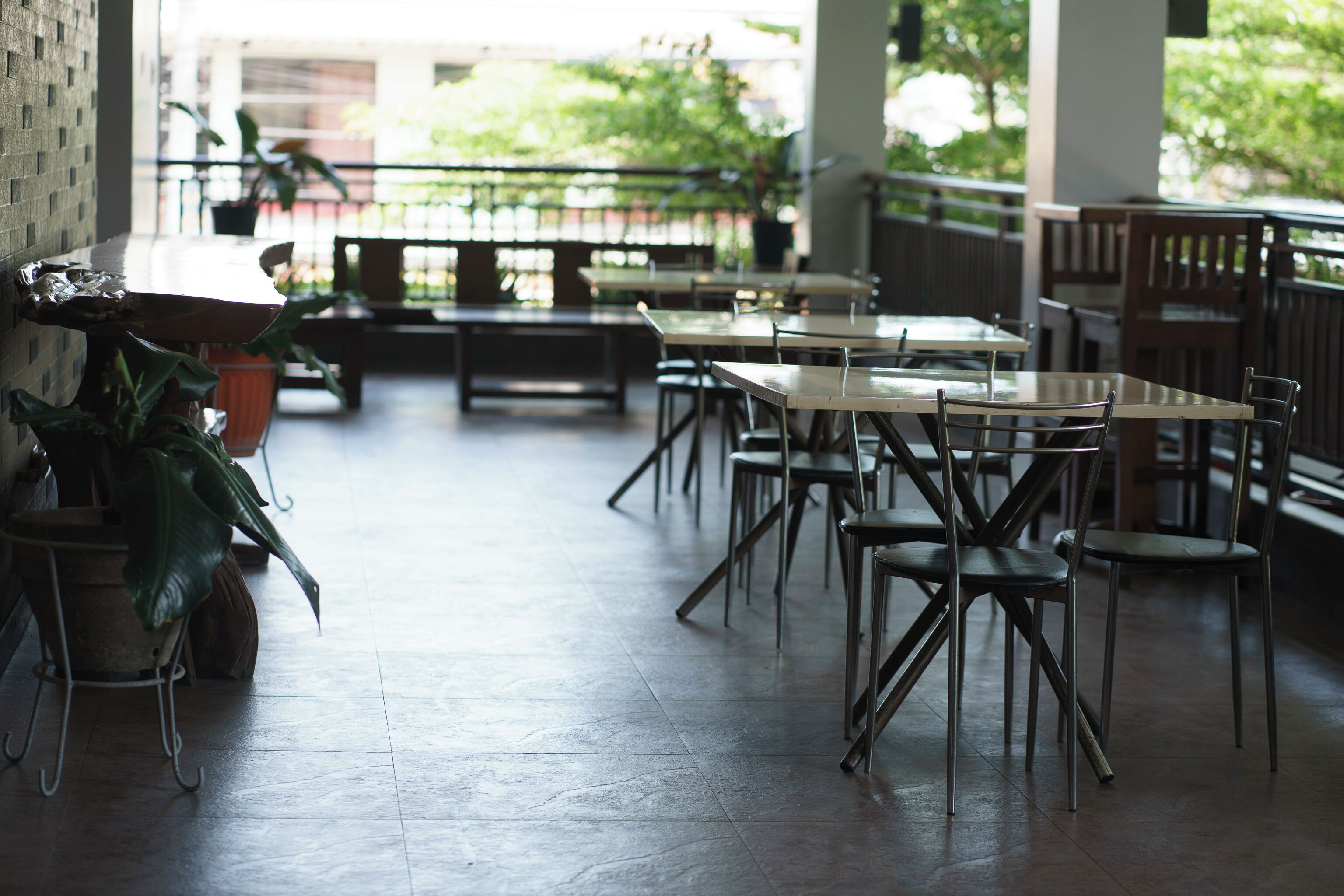 An outdoor cafe area with tables and chairs, potted plants, and a view of greenery.