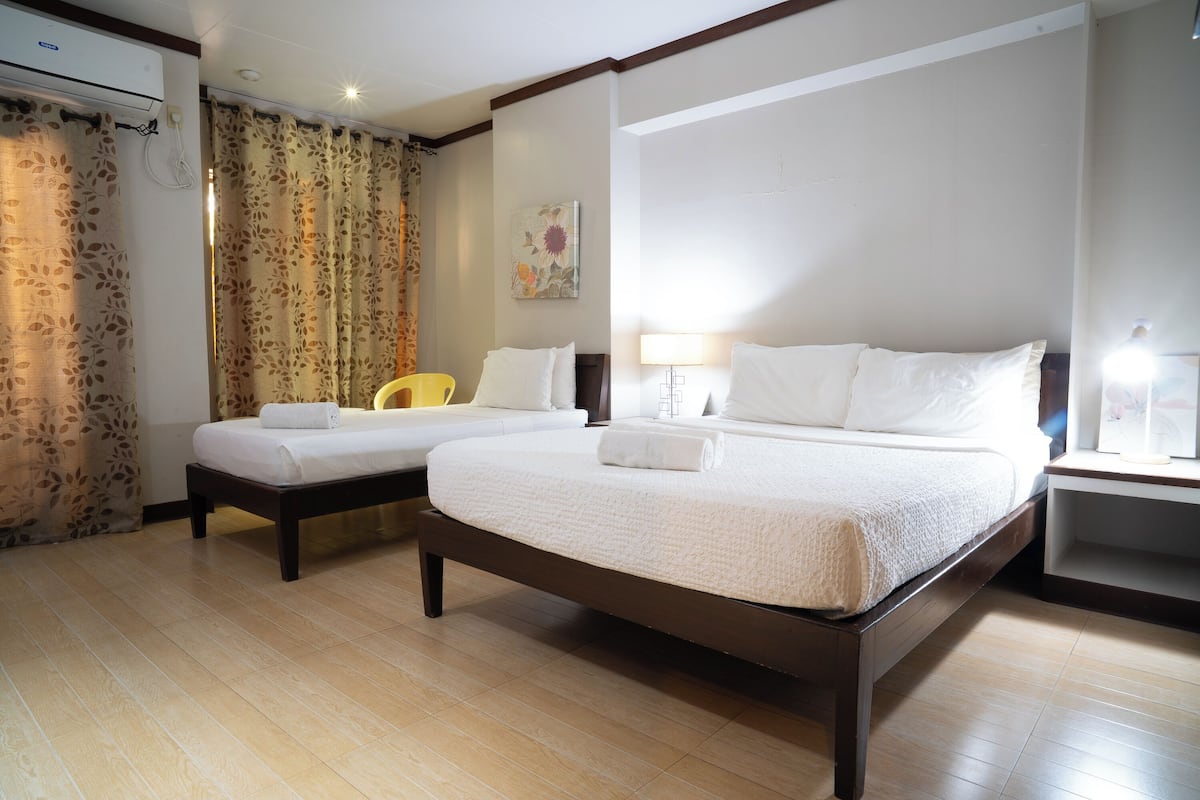 A hotel room with a large bed, two smaller beds, a chair, a nightstand, and a wall-mounted air conditioner.