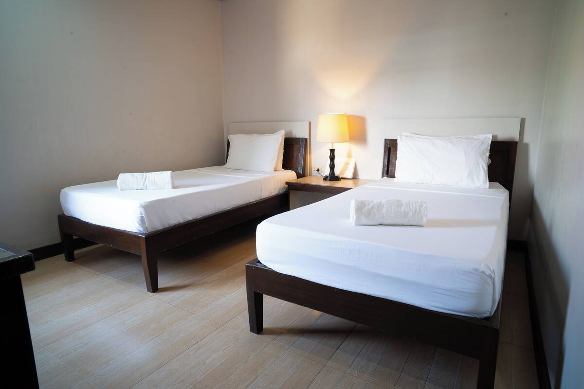 Two single beds with wooden frames and white linens, a nightstand with a lamp between them, and a tiled floor.