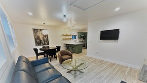 A modern living room with a black leather sofa, a glass coffee table, and a dining area with a round table and chairs. A kitchen with green cabinets and a flat-screen TV is visible in the background. - Outlaw Inn Unit 4.  Newly Remodeled large unit! 2 minutes walk to Main Street.  (Ouray)