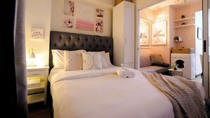 Un lit bien fait, avec du linge de lit blanc et des coussins décoratifs, une table de chevet avec une lampe, et un mur orné d’œuvres d’art encadrées.