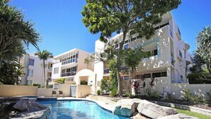 A swimming pool with clear blue water, surrounded by rocks and plants. There are modern buildings with balconies and large windows in the background. - Portsea North Apt 5  (Mooloolaba)