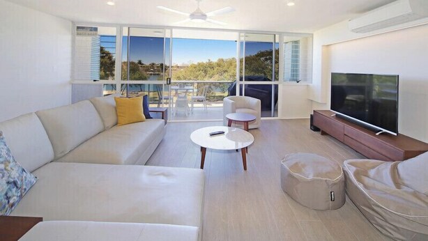 A modern living room with a sofa, a coffee table, and a TV. There is a balcony with a view of the outdoors. - Portsea North Apt 5  (Mooloolaba)