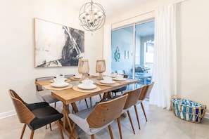 A dining area with a wooden table set for six, surrounded by chairs with wooden frames and cushioned seats. A chandelier hangs above, and a large abstract painting is on the wall.