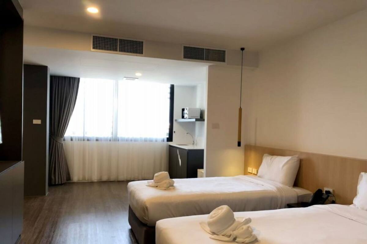 A hotel room with two beds, a desk, a chair, a window with curtains, and a ceiling vent.