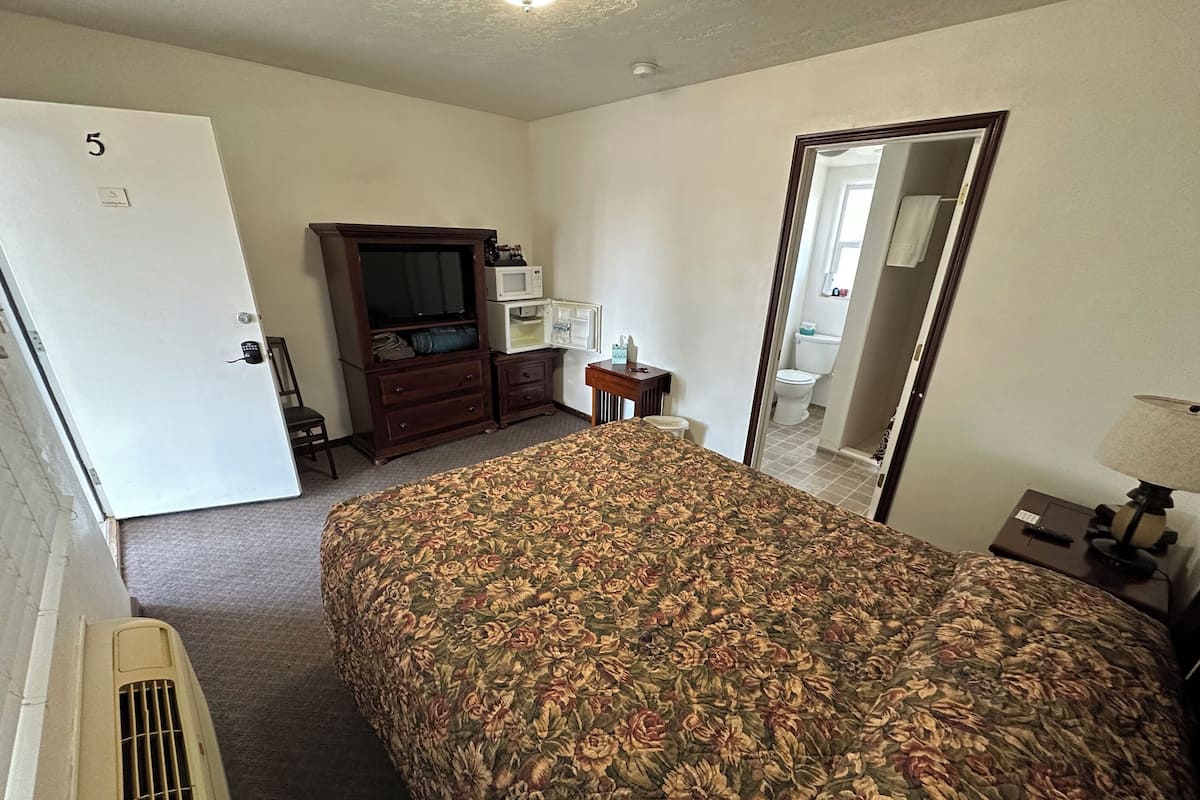 Ein Hotelzimmer mit einem blumengemusterten Bett, einer Holzkommode, einem Fernseher, einer Mikrowelle und einem durch eine offene Tür einsehbaren Badezimmer.