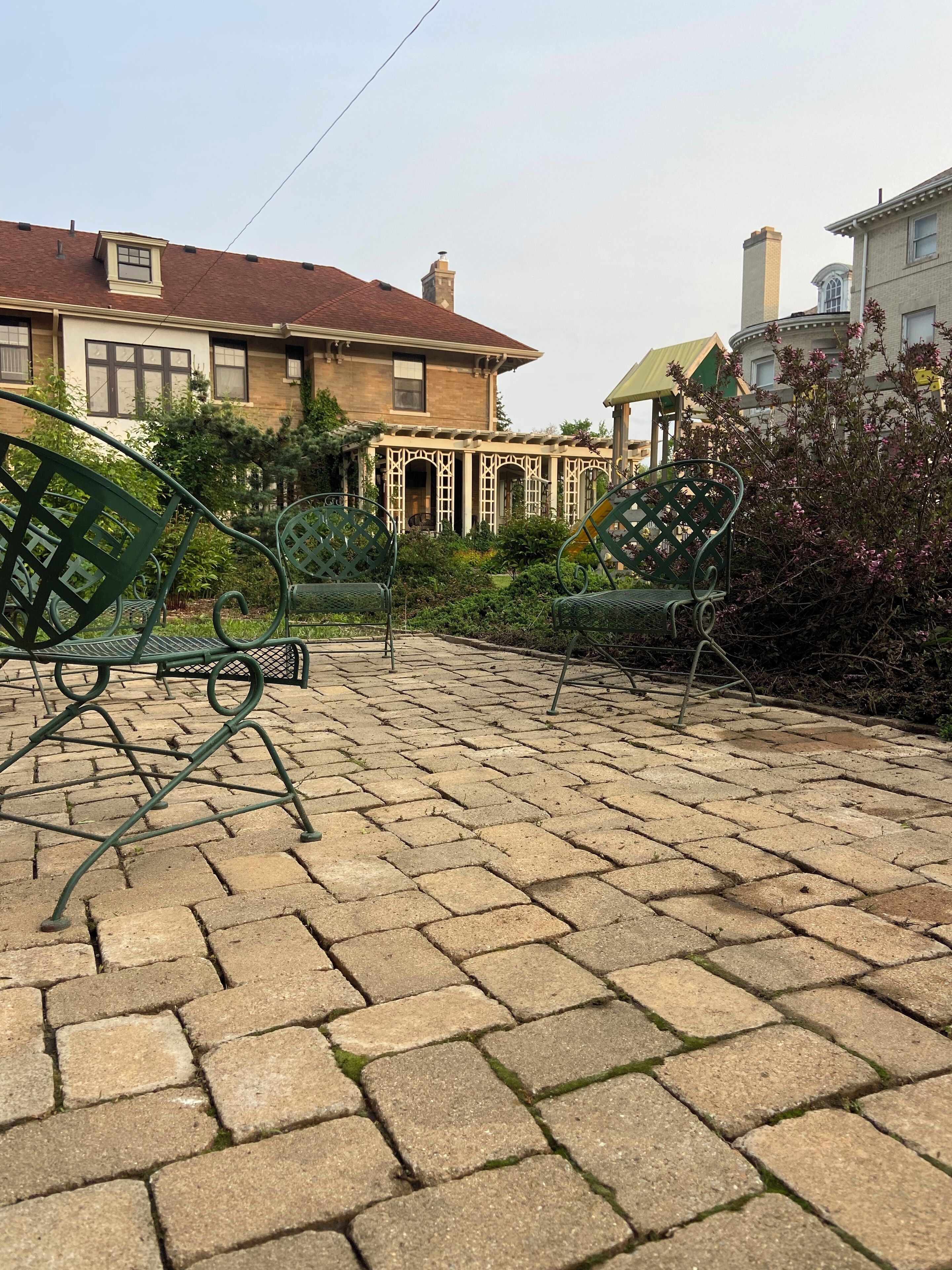 A paved patio with green metal chairs, a covered seating area, and a house with a red roof and beige walls.