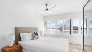 Un dormitorio con cama, mesita de noche, lámpara, ventilador de techo y vistas al exterior a través de amplias ventanas.