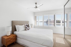 A bedroom with a bed, a nightstand, a lamp, a ceiling fan, and a view of the outside through large windows.