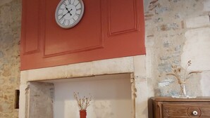 A room with a wooden floor, a fireplace with a clock above it, a small table with a vase, and a wooden cabinet.