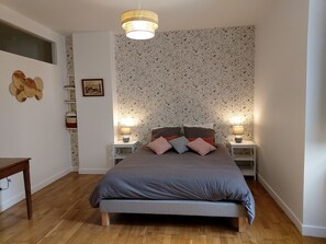 Une chambre à coucher avec un lit, deux tables de chevet avec des lampes, un plancher en bois, un mur orné d’un motif floral et une fenêtre.