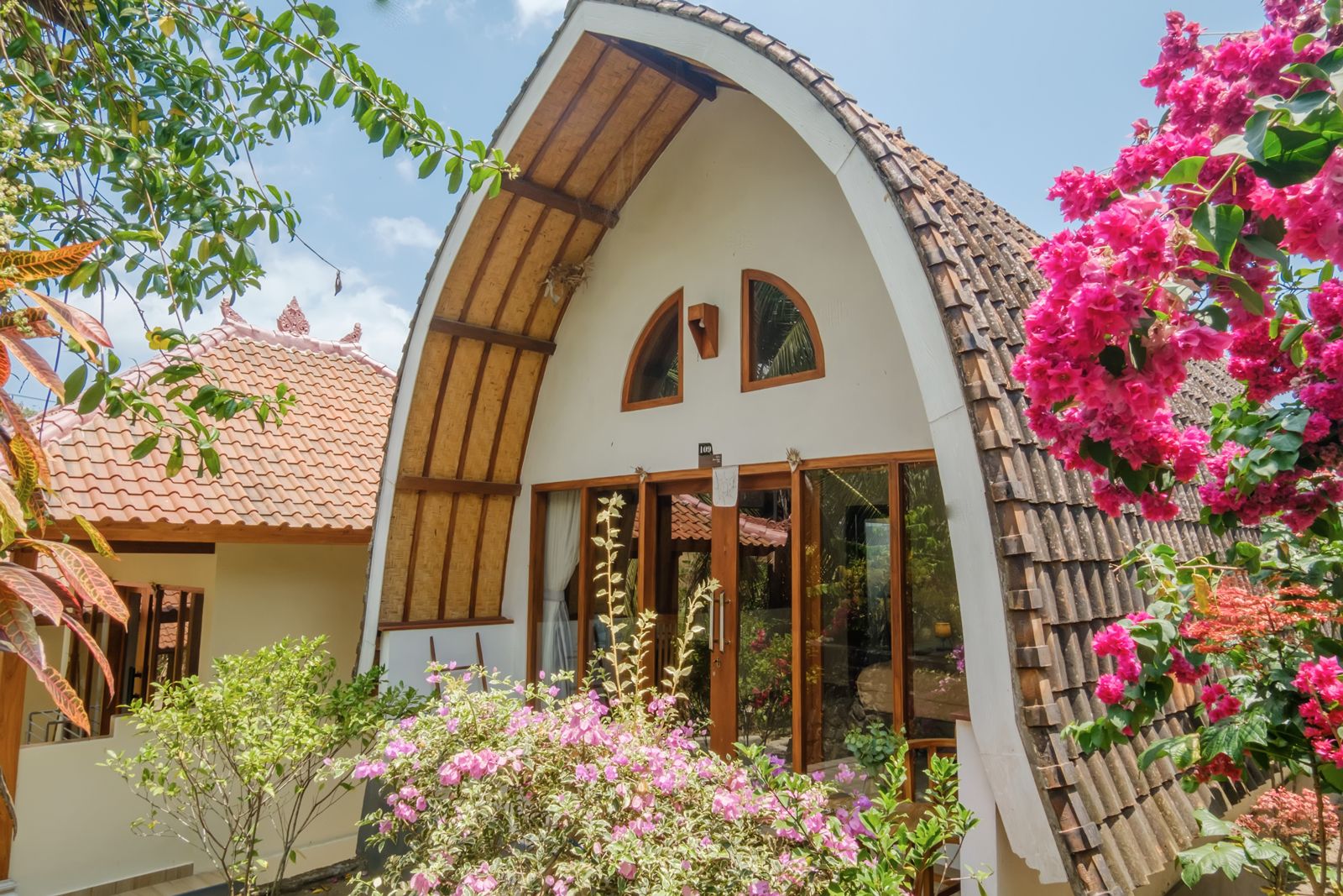Una casa con techo curvo, ventanas grandes y un jardín con flores rosadas.