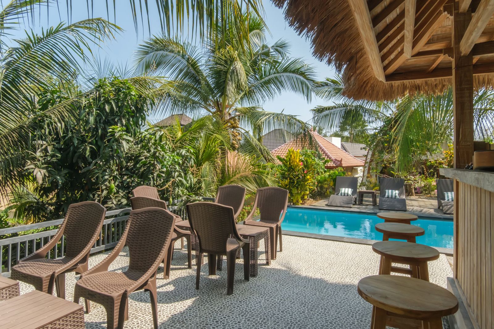 A poolside area with wicker chairs and tables, surrounded by palm trees and a clear view of a building in the background.
