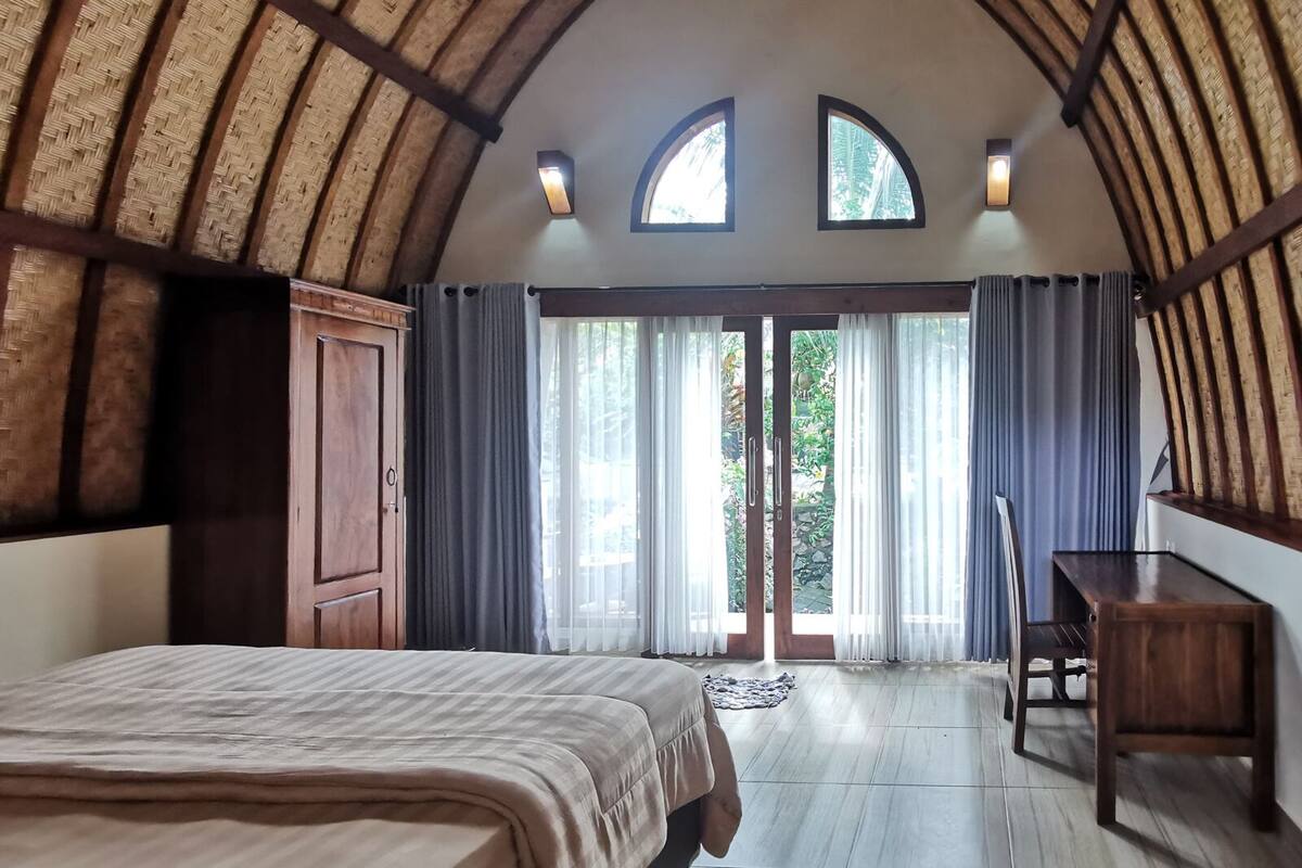 A bedroom with a large bed, a wooden desk, and a wardrobe. There are two windows and a door leading to a balcony.