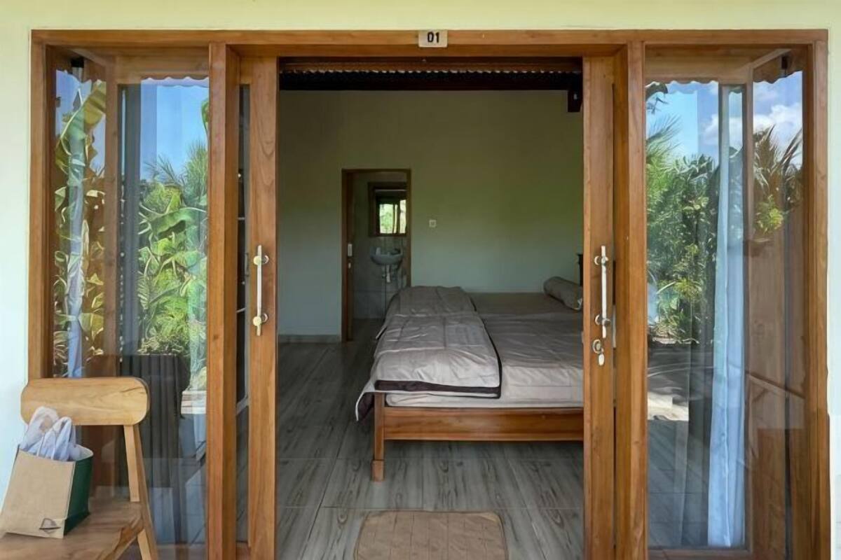 A room with a bed, a wooden door, and a view of greenery outside.
