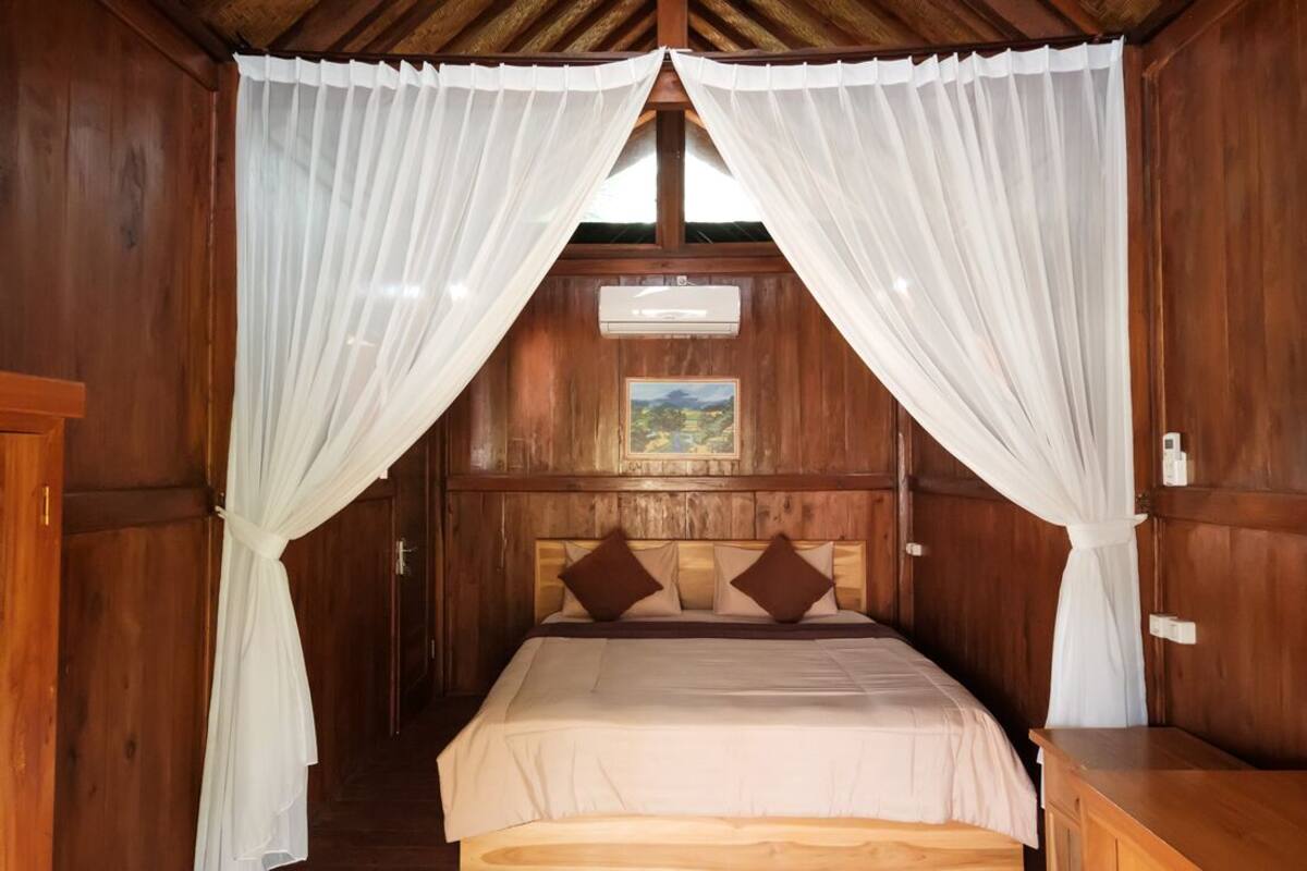 A wooden room with a bed, white curtains, and a painting on the wall.