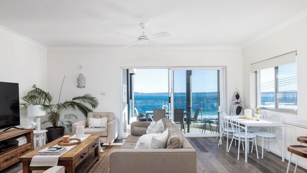 A modern living room with a sofa, coffee table, TV, and a balcony with a view of the ocean. - Bayview Towers 10 With Harbour Views (Nelson Bay)