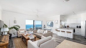 A modern living room with a sofa, coffee table, and bar stools. A kitchen with white cabinets and a large island is visible. - Bayview Towers 10 With Harbour Views (Nelson Bay)