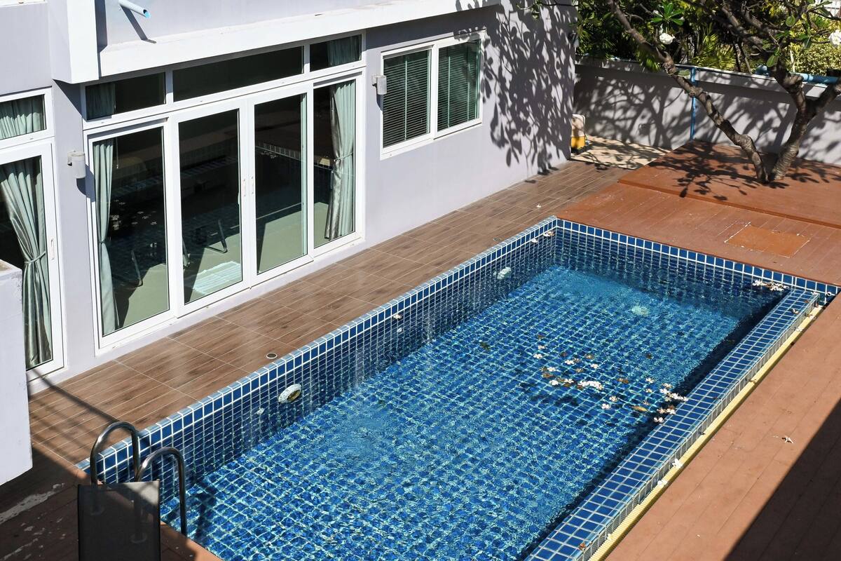 A rectangular swimming pool with blue tiles, surrounded by a wooden deck and a white building with large windows.