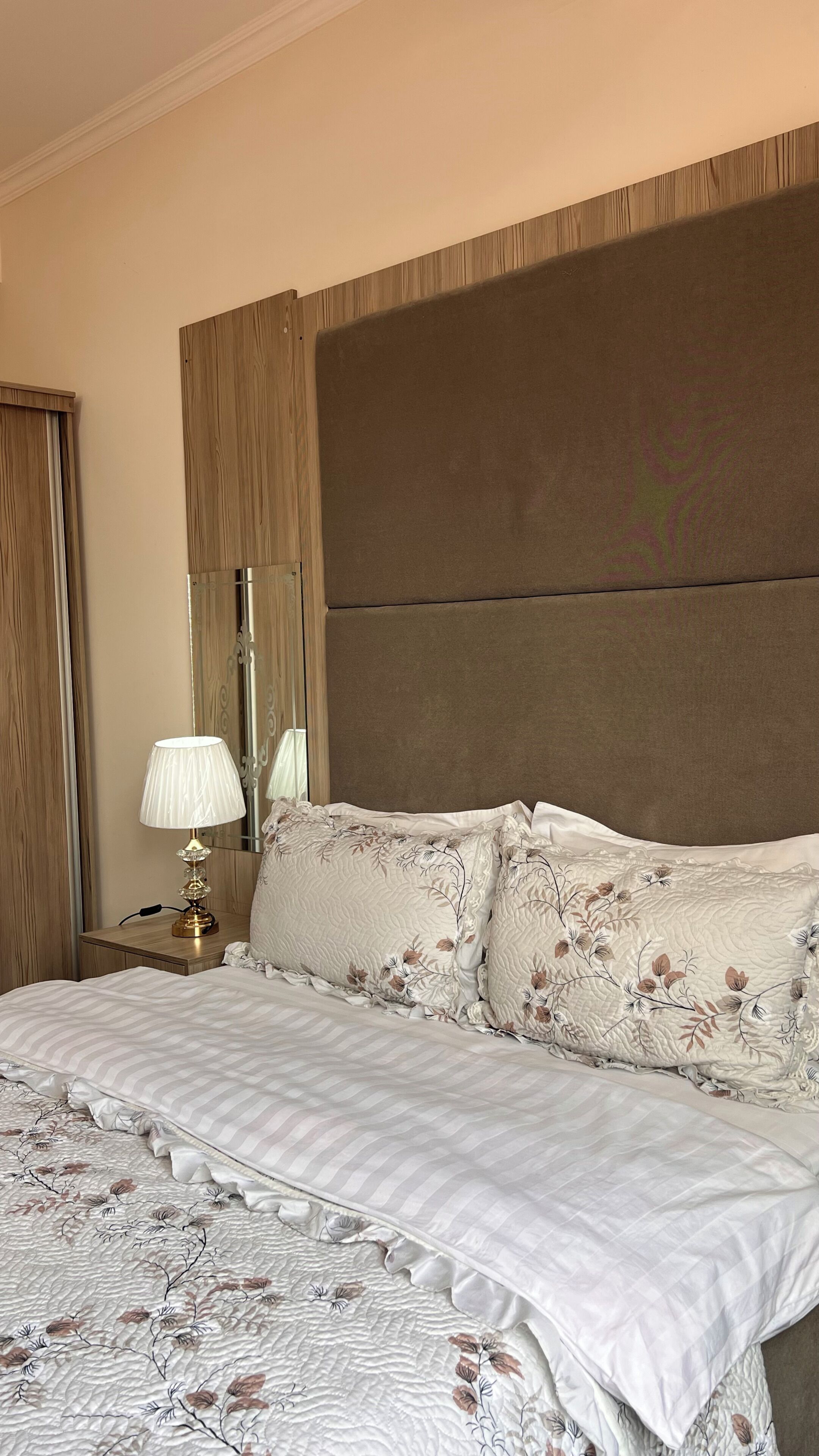 A bedroom with a large bed, a nightstand with a lamp, a mirror, and a wooden headboard.