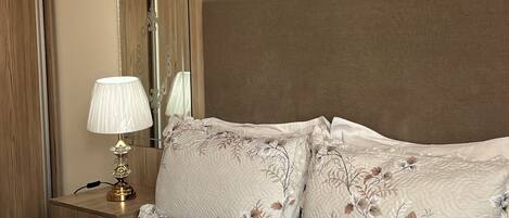 A bedroom with a large bed, a nightstand with a lamp, a mirror, and a wooden headboard.