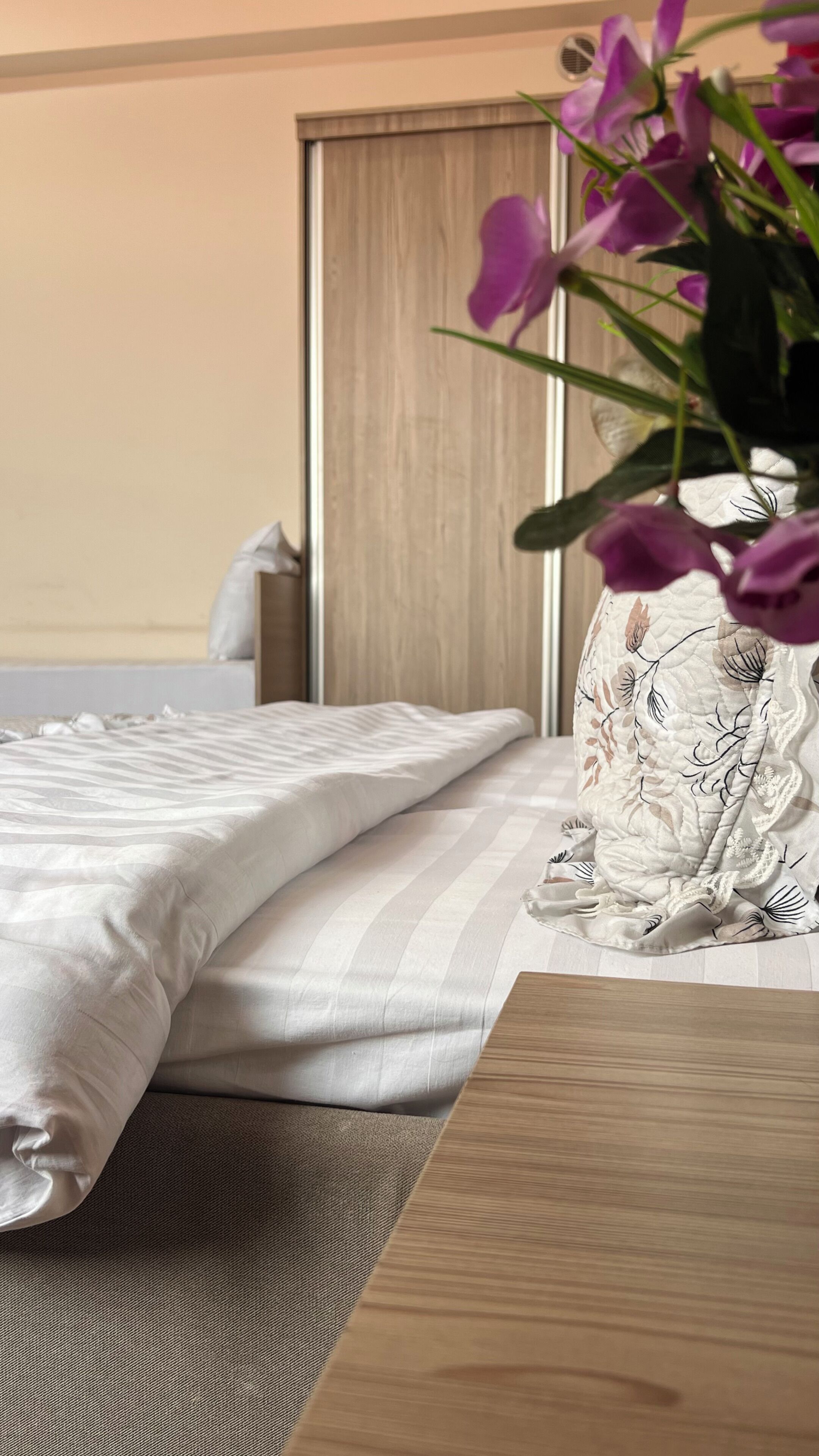 A neatly made bed with a striped bedspread, a wooden bedside table, and a vase with purple flowers.